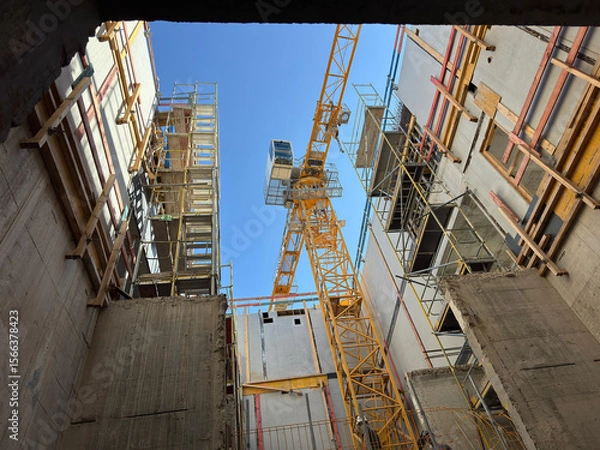 Fototapeta View looking up at a construction site with scaffolding and a yellow tower crane between partially built concrete structures under a clear blue sky