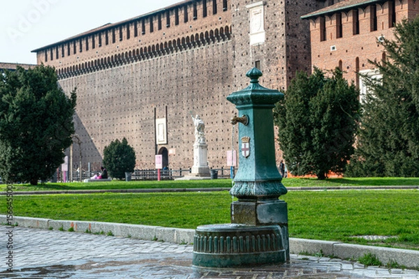 Obraz Antique green drinking fountain with the emblem of Milan in front of the historic Sforza Castle. The fountain stands on a cobblestone path surrounded by a well-kept park and lush green lawns.