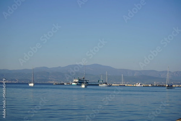 Obraz Sailboats gently gliding on calm waters at dawn near a tranquil harbor surrounded by distant mountains and clear skies