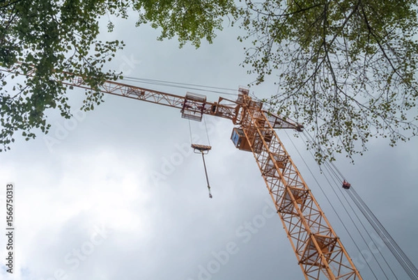 Fototapeta Equipment for the construction and lifting of bulky goods with construction materials. A tower crane on a construction site against a rainy sky. A construction high-rise crane and leaves on a tree.