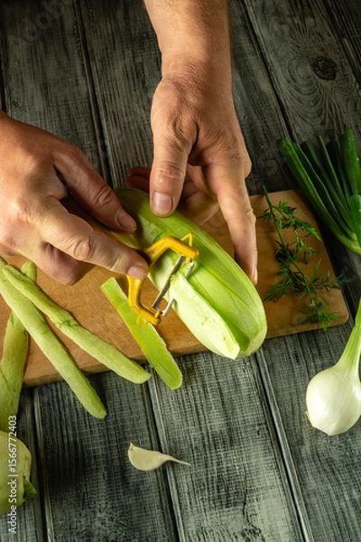 Fototapeta Fresh green vegetables marrow are being peeled on a wooden cutting board. Herbs and spring onions are arranged nearby, creating a vibrant and organic kitchen atmosphere