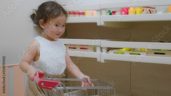 Fototapeta A young child explores a toy kitchen, engaging with colorful play food and a shopping cart in a bright playroom.
