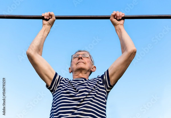 Fototapeta Portrait of an elderly man wearing glasses doing exercises on a horizontal bar at a sports ground. Staying active after retirement.