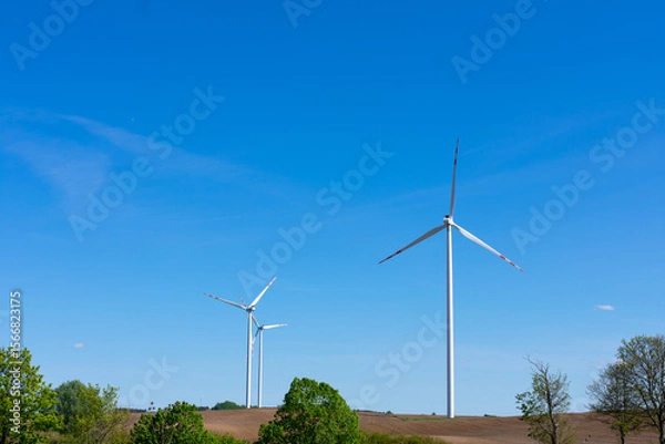Fototapeta Modern wind turbines generating renewable energy on a clear day, surrounded by fields and trees, symbolizing sustainable and clean power solutions.