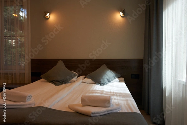 Fototapeta A fragment of a hotel room. A double bed with over-bed lamps. Mountain pines are reflected in the glass of the door.