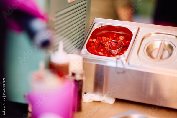 Fototapeta Close-up of a stainless steel soup warmer with a fruit-filled, red-colored beverage or sauce, accompanied by condiment bottles, set on a wooden table with a blurred background