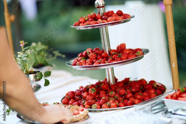 Fototapeta Outdoor three-tiered silver tray overflowing with ripe strawberries, surrounded by spring flowers and treats, set on a white tablecloth in lush garden sunlight, evoking a festive summer atmosphere