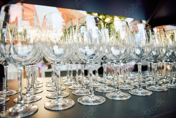 Fototapeta Elegant row of empty wine glasses on dark table outdoors, with sunlight reflecting off the glass surfaces, evoking a luxurious atmosphere for an event or celebration