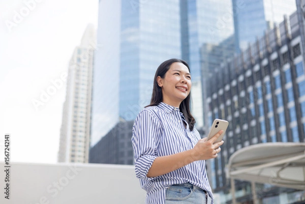 Fototapeta close up manager businesswoman hold smartphone and looking away through while stand outside office building tower for better vision and thinking about new business project for startup
