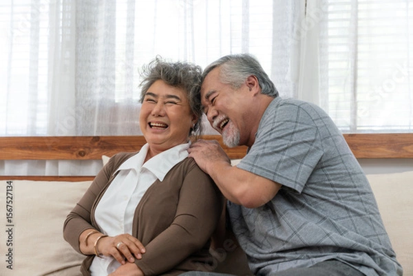 Fototapeta Asian elderly couple enjoying joyful moment together at home, smiling and laughing while sitting closely on a cozy sofa