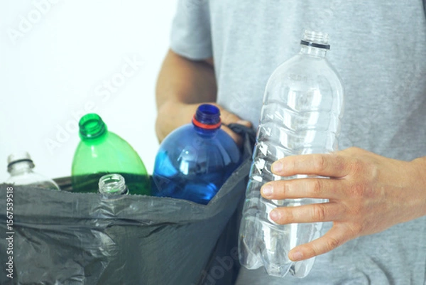 Fototapeta Everyday eco habit: sorting packaging for recycling. Focus on conscious living: a man holds a used transparent plastic bottle in one hand and a full garbage bag in the other. Waste separation