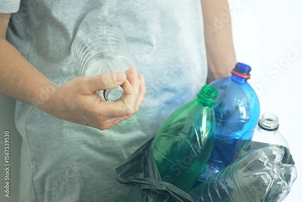 Fototapeta Sorting plastic waste at home: а man puts empty bottles without caps in a black garbage bag, symbolizing environmental care and household recycling habits. Eco responsibility and sustainable lifestyle