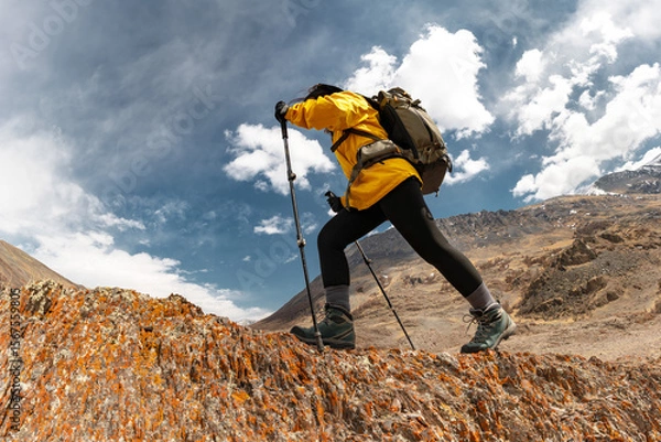 Obraz Girl trekker in hiking gear climbs a rock against the background of blue sky with clouds and rocky Altai mountains. Low angle bottom view. Adventure energy, endurance, solo hike
