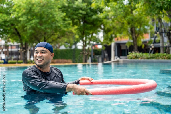 Obraz A smiling person in a swim cap and rash guard is in a pool, holding a pink swim noodle in an arc above their head. This image depicts aquatic exercise and joyful water recreation.