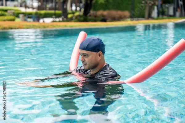 Obraz A smiling person in a swim cap and rash guard is in a pool, holding a pink swim noodle in an arc above their head. This image depicts aquatic exercise and joyful water recreation.