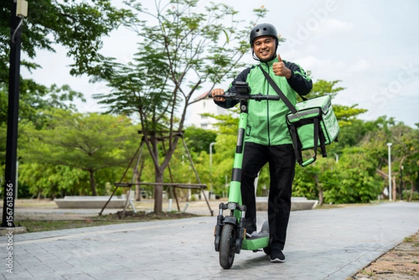 Obraz Rider food delivery man wearing green uniform and helmet ride the EV scooter delivery food service to customer. Happy delivery man with green backpack shipping of goods to customers by scooter