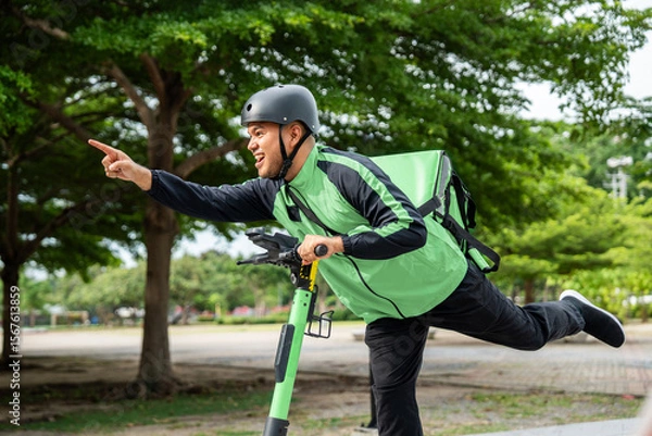 Obraz Rider food delivery man wearing green uniform and helmet ride the EV scooter delivery food service to customer. Happy delivery man with green backpack shipping of goods to customers by scooter