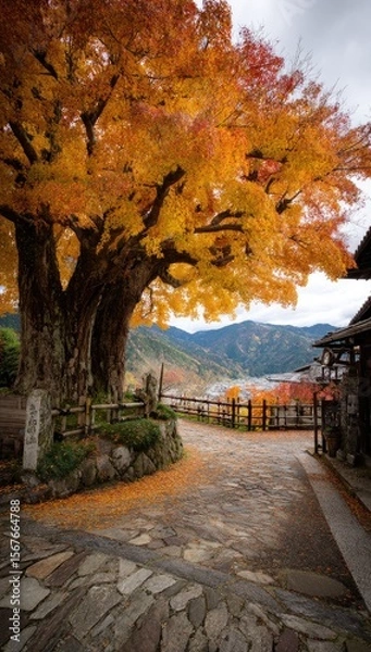 Obraz Autumnal path beneath a vibrant maple tree, leading to a mountain village