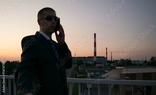 Fototapeta A silhouette of a man in a suit and tie, talking on the phone, against the background of a factory with pipes 