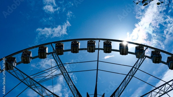 Fototapeta Ferris wheel photographed from below against a blue sky with sun flare and scattered clouds.