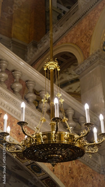 Fototapeta Golden chandelier with candle-like bulbs hanging in an opulent classical interior with stone railings.