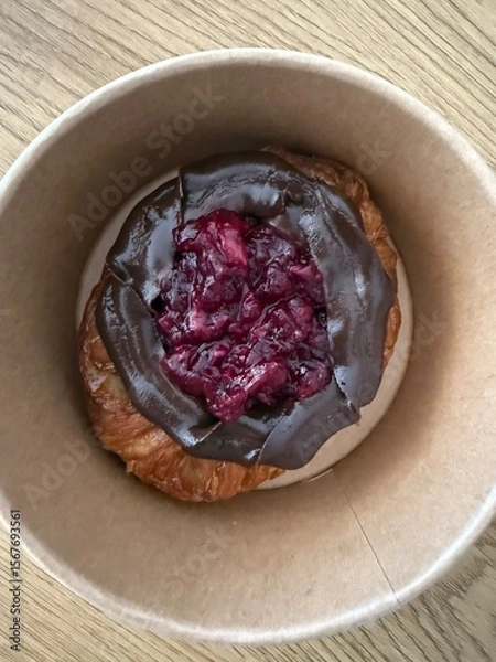 Fototapeta A close-up of a croissant in a paper bowl, topped with chocolate spread and a cherry filling, placed on a wooden surface.