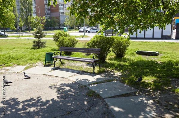 Fototapeta Rest area with bench surrounded by trees and ornamental shrubs in Kyiv, Europe. Place to rest in the city at spring