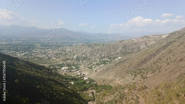 Fototapeta High-angle view of a serene mountain valley town, nestled amongst arid hills and distant mountains under a partly cloudy sky.