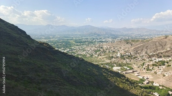 Fototapeta Serene Mountain Valley Landscape: Panoramic View of Village Nestled Amongst Lush Greenery and Majestic Mountains Under a Partly Cloudy Sky.