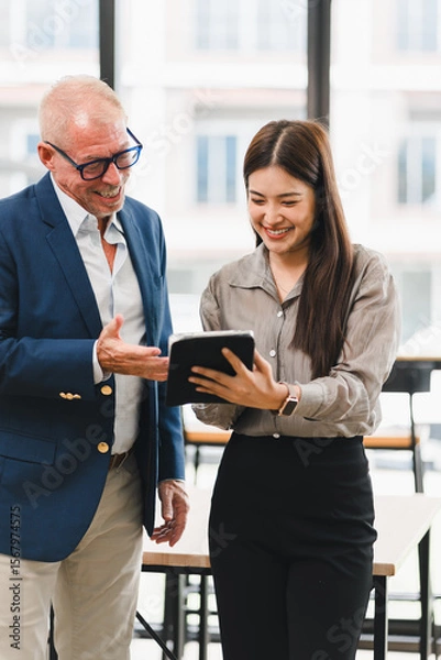 Fototapeta Smiling businesswoman showing digital tablet to senior businessman in modern office, teamwork and collaboration concept, both appear happy and engaged in discussion