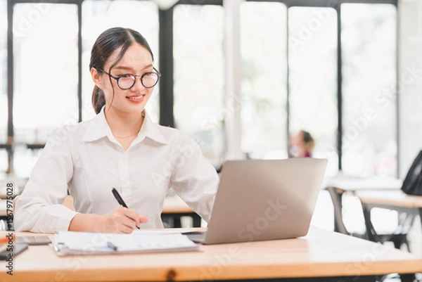 Obraz Young Asian woman, wearing glasses and white shirt, smiling while working on laptop and taking notes in modern bright office, professional and focused atmosphere