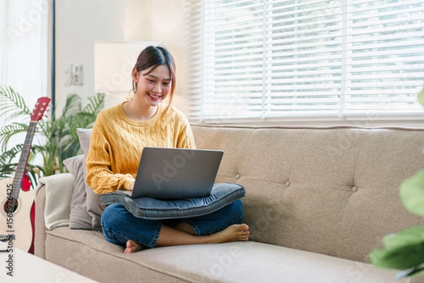 Fototapeta Young woman sitting cross legged on sofa with laptop, smiling and working in bright living room with natural light, cozy atmosphere, casual home workspace