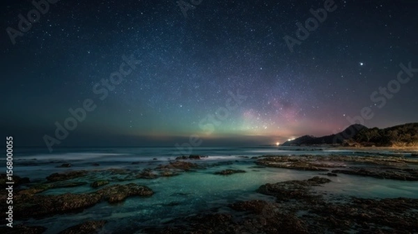 Fototapeta Starry night sky illuminates a rocky shoreline with gentle waves and distant mountains a tranquil
