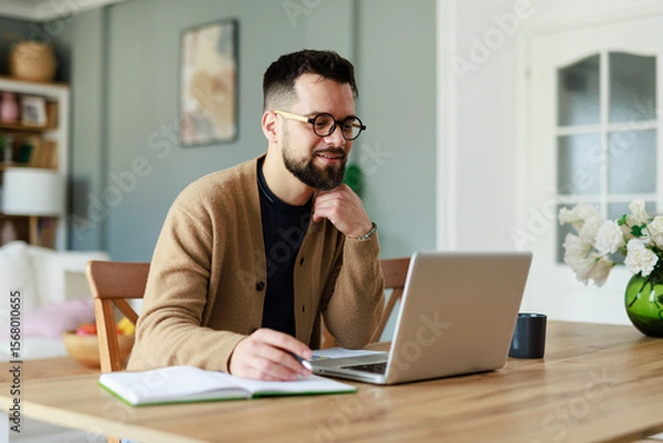 Fototapeta Young Caucasian Man Working Remotely in a Cozy Home Office Setting with Laptop and Notebook