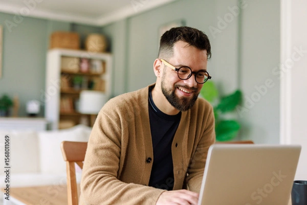 Fototapeta Smiling Young Caucasian Man in Glasses Working Remotely from Home in a Warm and Cozy Living Room