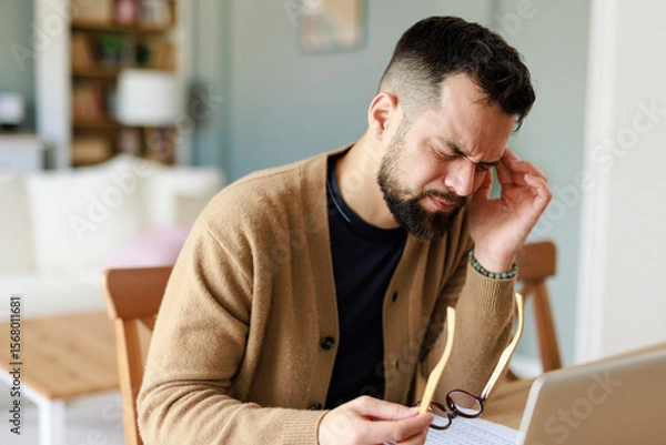 Fototapeta Young Caucasian Man in Pain Working from Home Holding Glasses in a Comfortable Indoor Environment