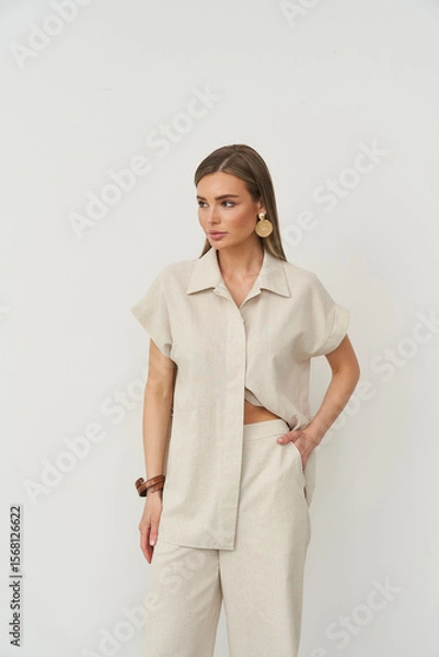 Fototapeta Model in beige short-sleeve shirt and wide-leg pants against plain backdrop. Fashion-forward look with minimalistic style, accessorized with large earrings and a bracelet