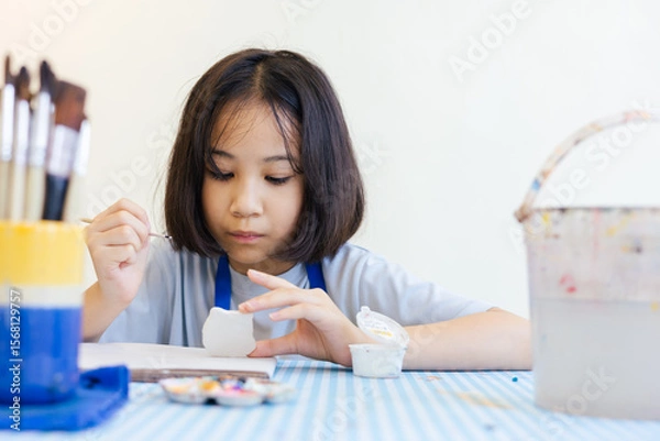 Obraz Young Asian girl focuses on painting a clay object, with her hand and brush with selective focus shows creativity and calm concentration for kid learning activity  via art and development in child.