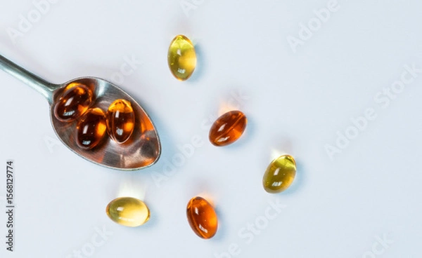 Obraz A close-up, top-view image of golden fish oil capsules on a silver spoon, isolated on a clean white background. This flat lay composition represents the concept of daily nutritional supplements.
