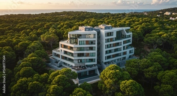 Fototapeta Aerial view reveals a modern white building surrounded by lush green forest, with the sea visible in the distance
