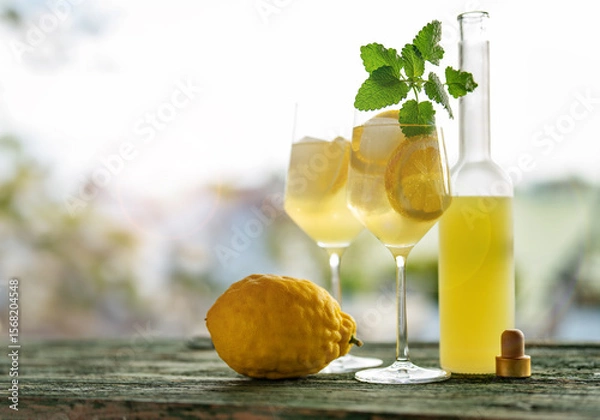 Obraz Limoncello liqueur with ice cubes in glasses and a ripe lemon on rustic wooden table.Traditional italian refreshing summer drink. 