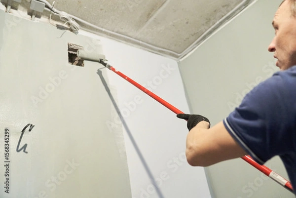 Fototapeta A man paints concrete walls with a paint roller. Gray paint creates a cozy atmosphere in the room. High quality photo