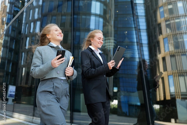 Fototapeta Business professionals in smart attire engage with devices while walking in a modern urban setting during a sunny day