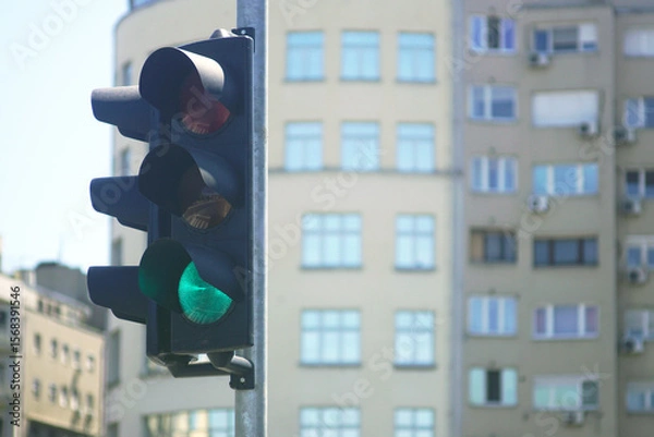Fototapeta Urban traffic light showing green signal at city intersection with blurred modern residential buildings in background. Concept of safe driving, road regulation, navigation and transport infrastructure