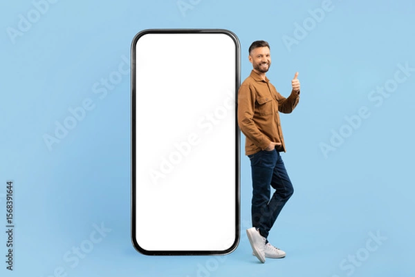 Fototapeta Cheerful young man standing with thumbs up next to big smartphone mockup. Ideal for app marketing, mobile UI design, or tech startup concepts on blue background, collage