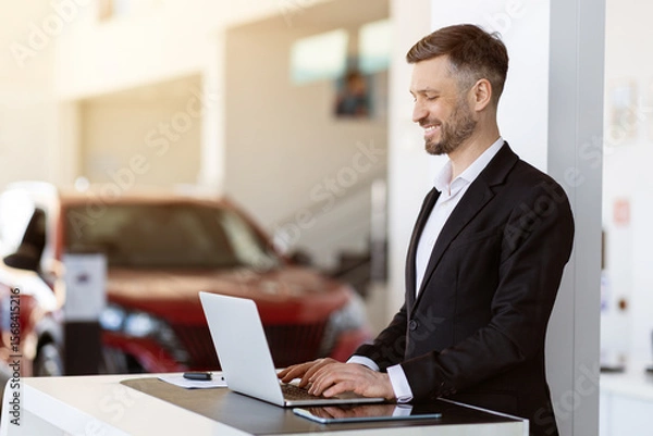 Fototapeta Professional car salesman working at laptop in dealership showroom. Handsome man preparing documents or checking inventory with a smile in bright, elegant workspace, copy space