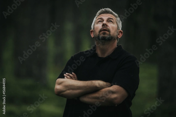 Fototapeta A man with gray hair and a beard stands with his arms crossed, looking up into a shady forest. He is wearing a black shirt, and his mood is contemplative and calm.