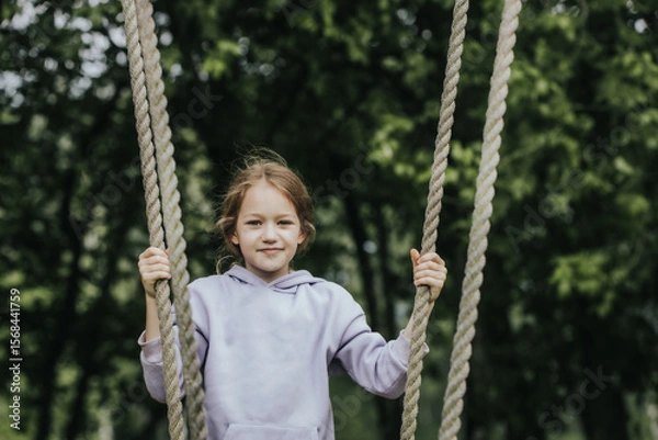 Fototapeta A cheerful girl in a lavender hoodie holds the ropes of a wooden swing in a park surrounded by deciduous trees and smiles directly at the camera. Natural daylight and greenery in the background.