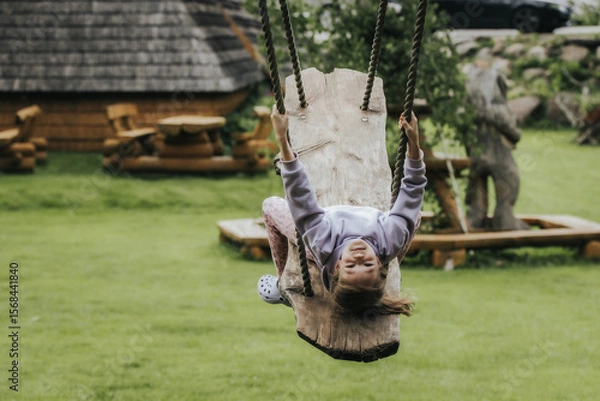 Fototapeta A happy girl in a lavender sweatshirt swings upside down on a rustic wooden swing in a grassy yard with wooden decor and parked cars in the background.