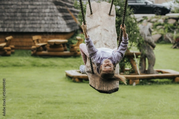 Fototapeta A happy girl in a lavender sweatshirt swings upside down on a rustic wooden swing in a grassy yard with wooden decor and parked cars in the background.